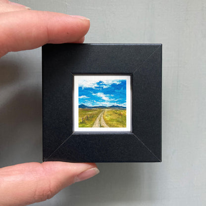 A person's hand holding a small black picture frame containing a miniature landscape painting depicting a winding road leading to a blue sky with clouds.