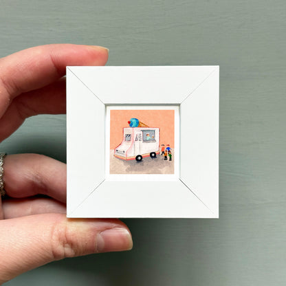 A hand is holding a small white picture frame containing an illustration of an ice cream truck.