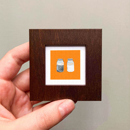 A hand is holding a small square frame containing an illustration of two salt and pepper shakers.