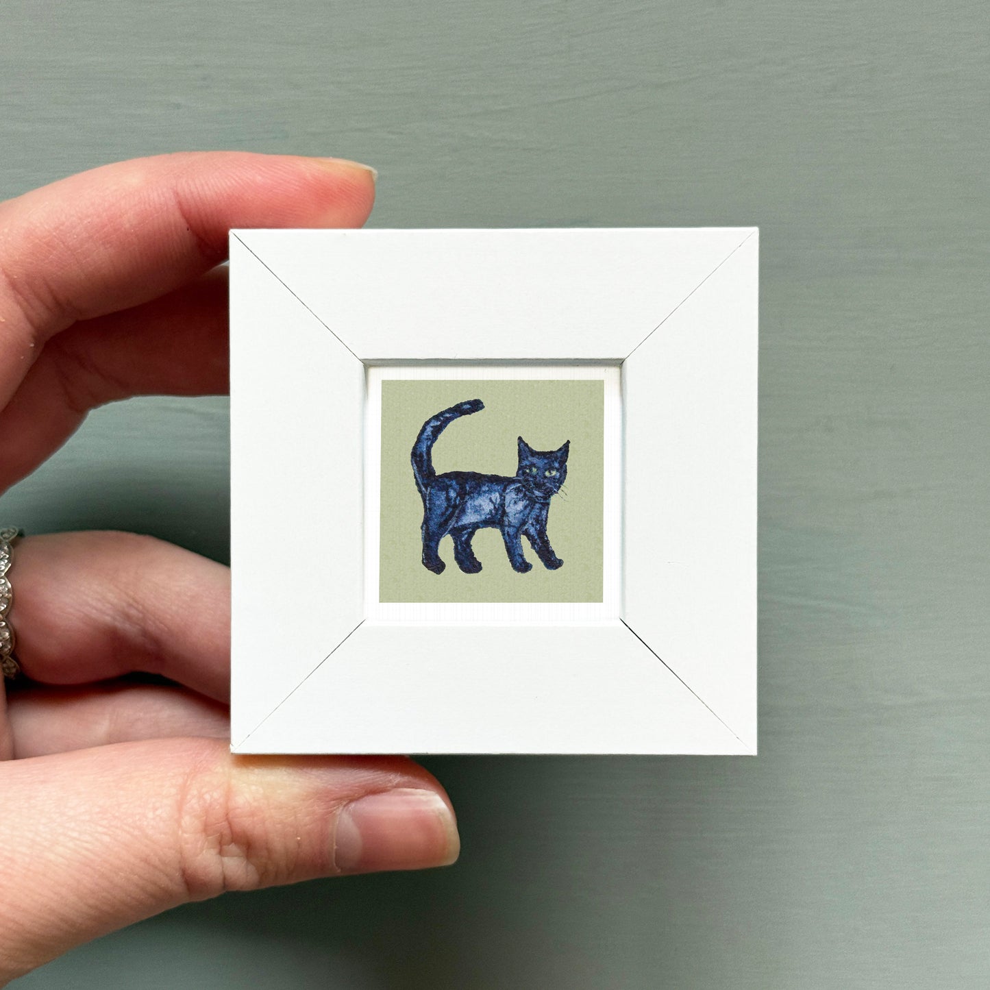 A hand is holding a small white square frame containing a black cat illustration.