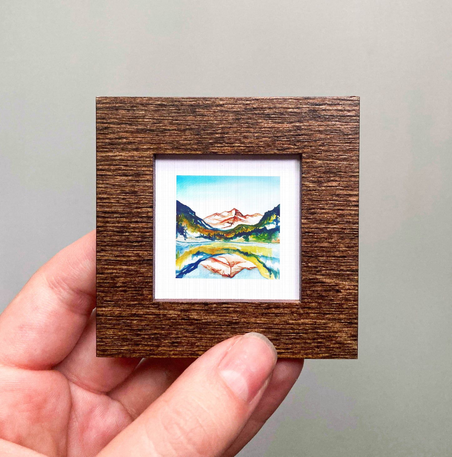 A hand is holding a small, square-shaped wooden frame containing a painting of a mountain landscape.