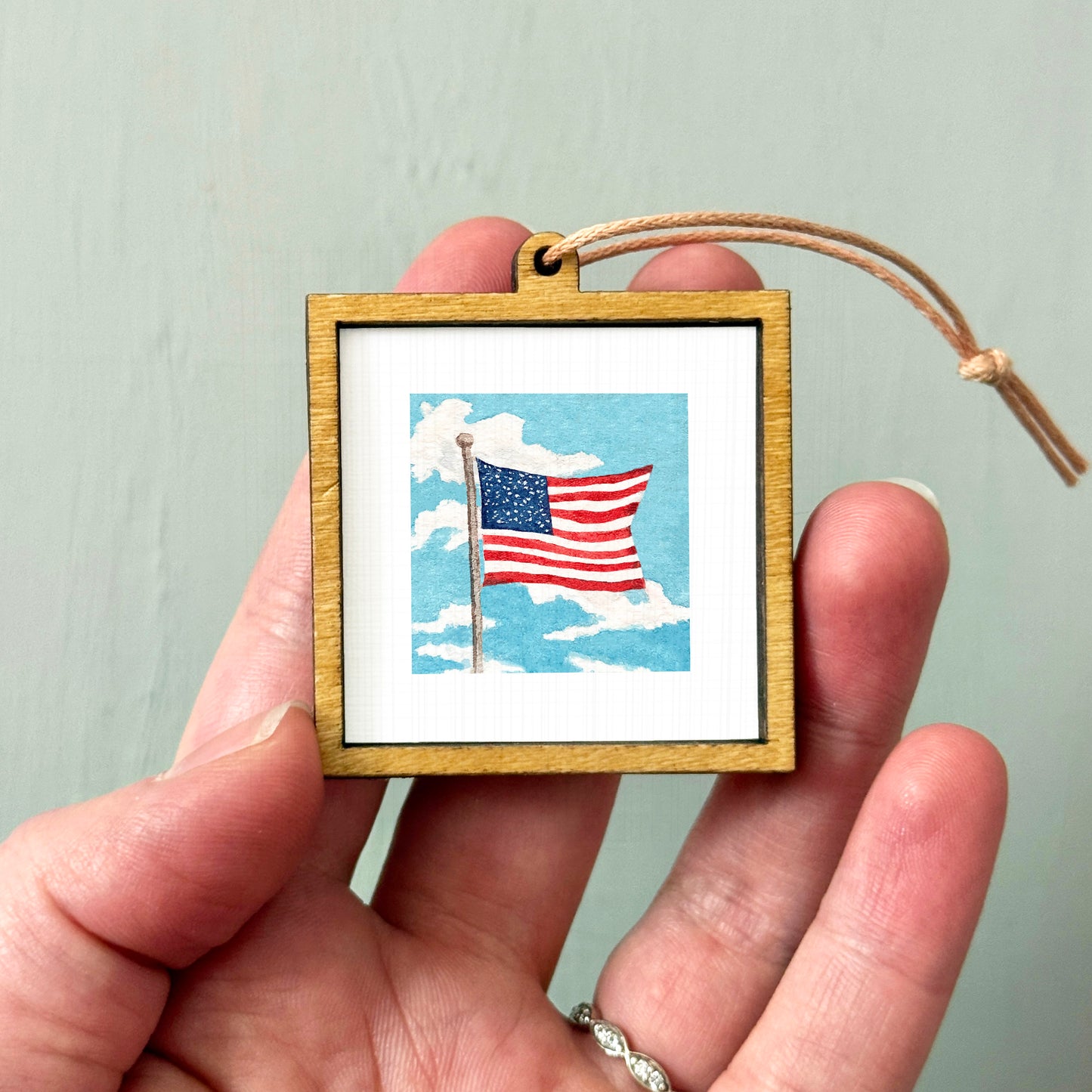 A hand holding a small wooden frame containing a picture of the American flag against a cloudy sky background.