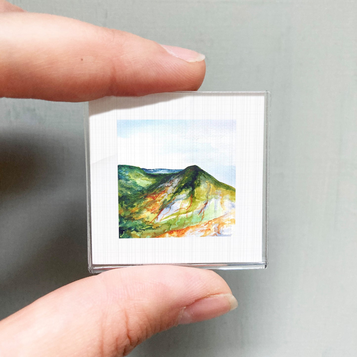 A person's hand holding a small, square-shaped picture of a mountain landscape.