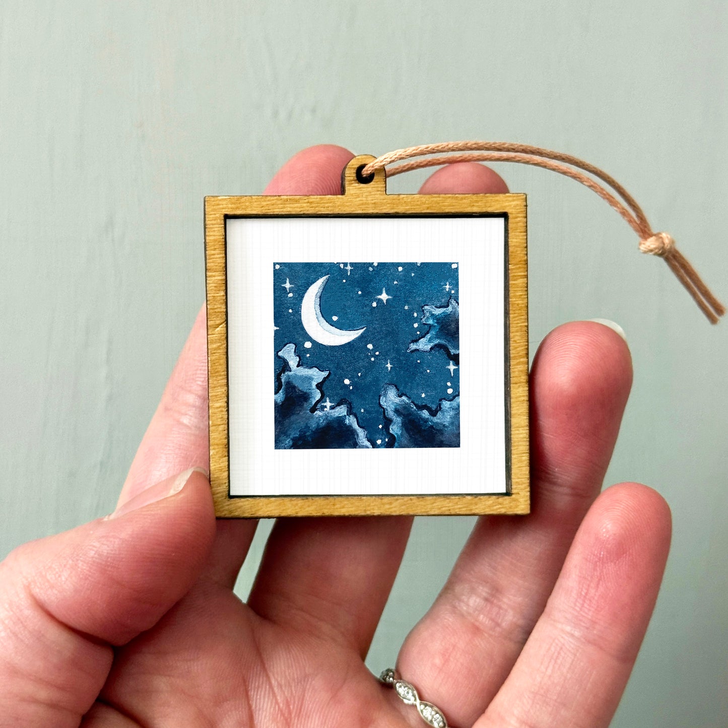 A hand holding a small wooden frame containing a watercolor painting of a crescent moon and starry night sky.