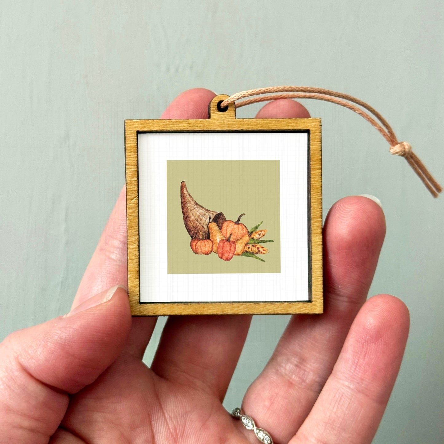 A person's hand holding a small wooden frame containing a framed image of a Thanksgiving-themed illustration.