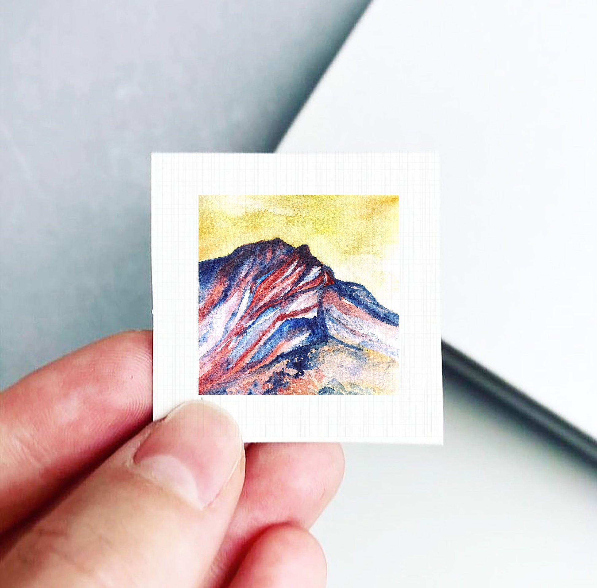 A hand is holding a small square piece of paper with a watercolor painting of a mountain landscape on it.