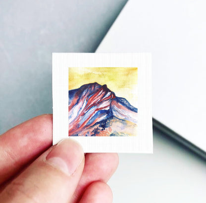 A hand is holding a small square piece of paper with a watercolor painting of a mountain landscape on it.