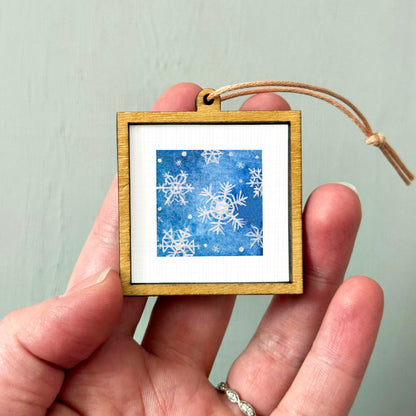 A hand holding a small wooden frame containing a picture of snowflakes against a blue background.