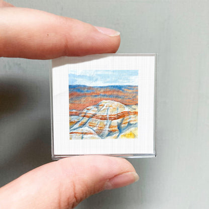 A person's hand holding a small, square-shaped picture of a landscape with mountains and a blue sky.