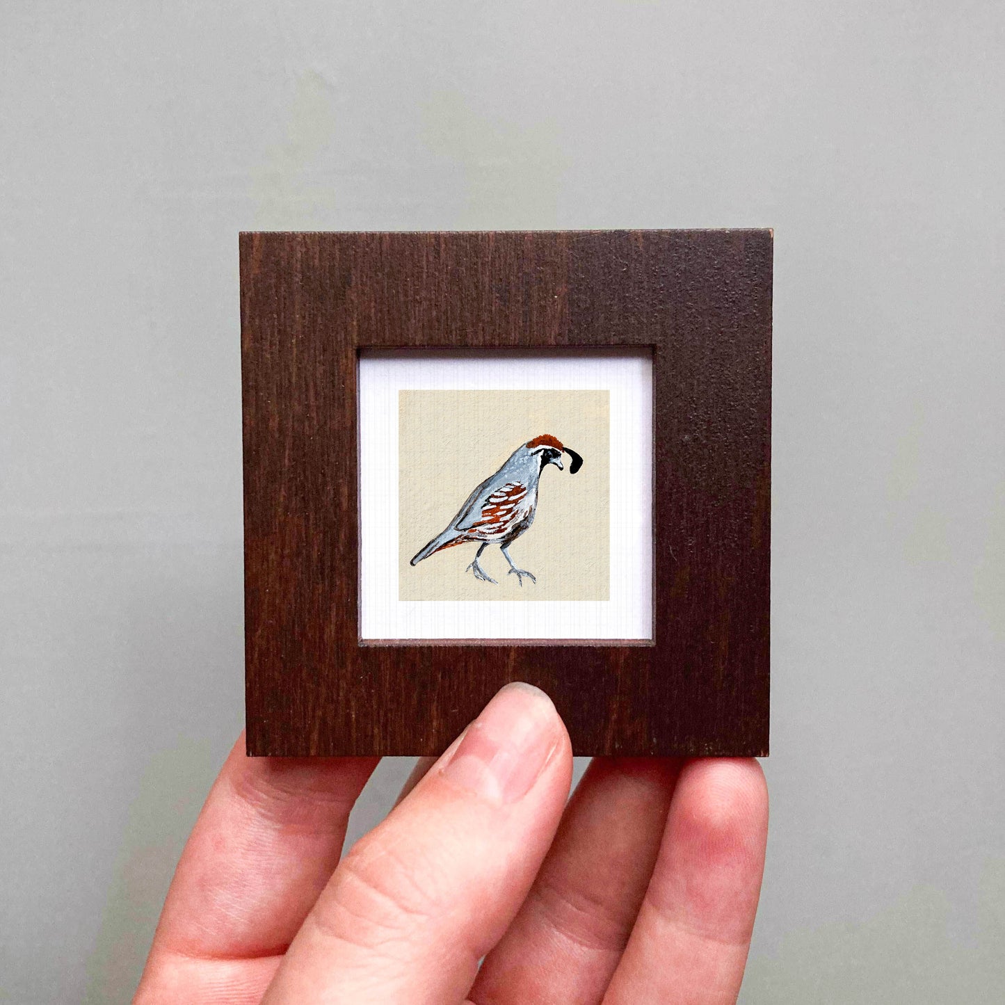 A person's hand is holding a small wooden frame containing a framed illustration of a bird.