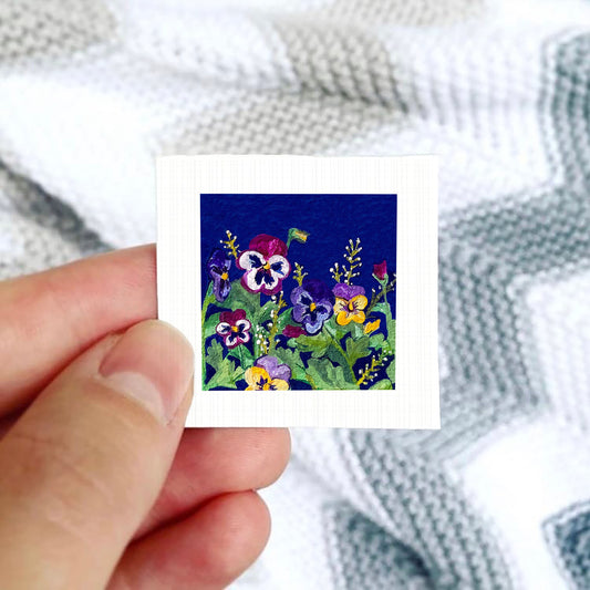 A hand is holding a small square card with a vibrant floral design featuring colorful pansies and other flowers against a blue background.