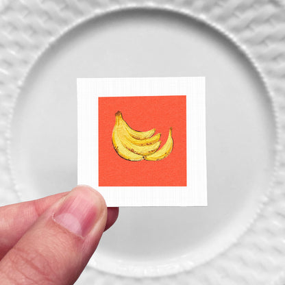 A hand is holding a small square with a vibrant red background and a depiction of bananas.