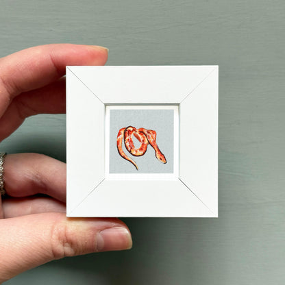 A hand is holding a small white square frame containing a painting of a red snake.