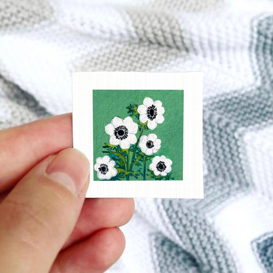 A hand is holding a small square card with a painting of white flowers against a green background.