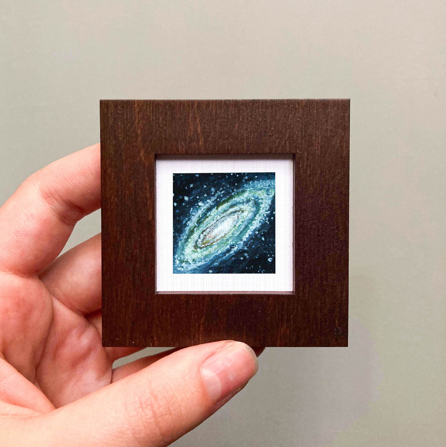 A hand is holding a small wooden frame containing a framed image of a spiral galaxy.