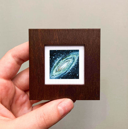 A hand is holding a small wooden frame containing a framed image of a spiral galaxy.