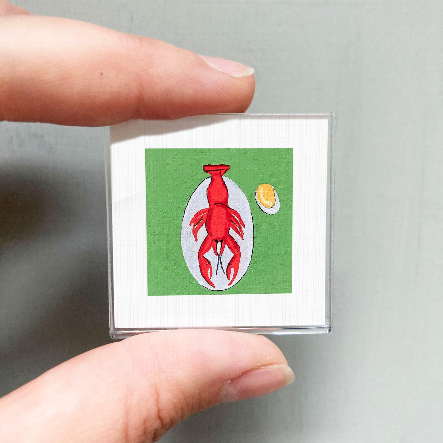 A person's hand is holding a small square picture depicting a red lobster on a green background.