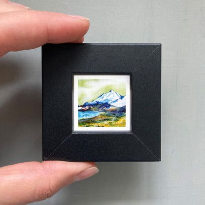 A person's hand holding a small, square-shaped black frame containing a watercolor painting of a mountain landscape.