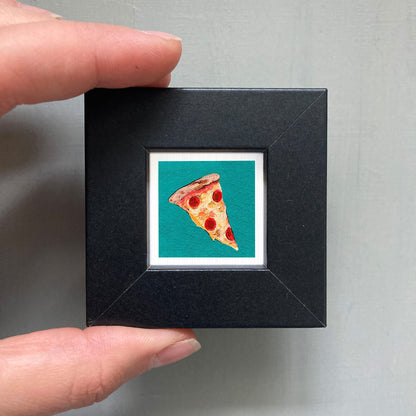 A hand is holding a small black frame containing a framed image of a slice of pizza.