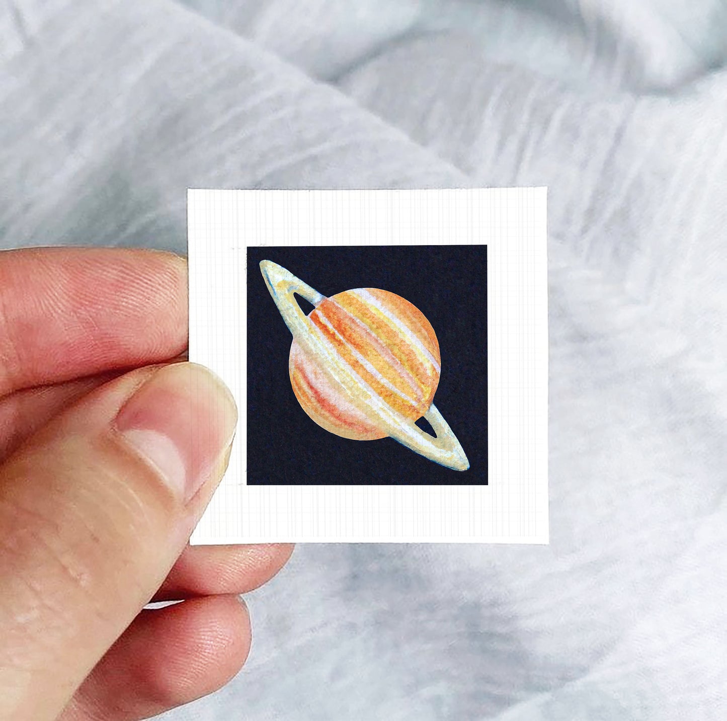 A hand is holding a small square-shaped paper with a depiction of a planet, specifically the planet Saturn, on it.