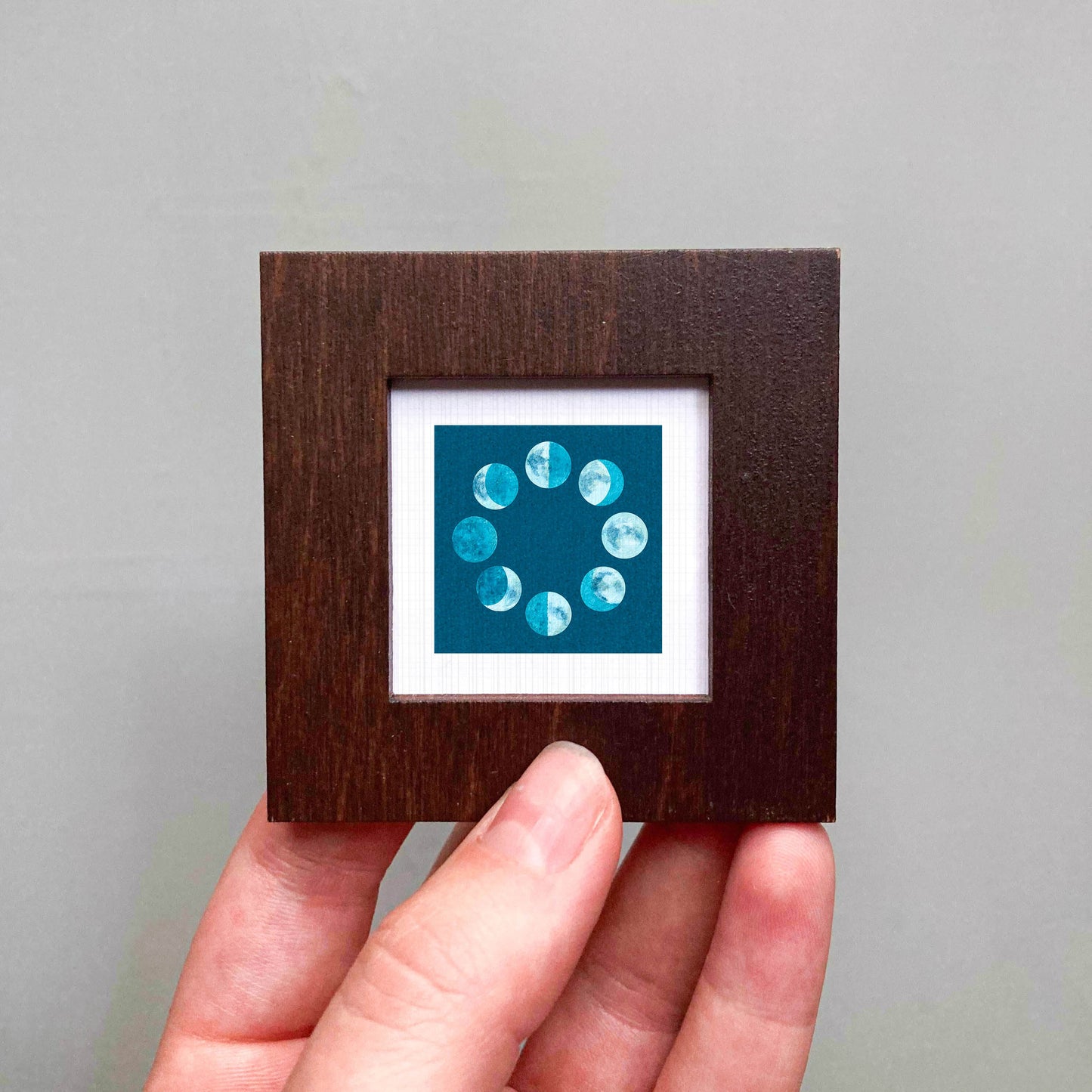 A hand is holding a small wooden frame containing a framed image of the phases of the moon.