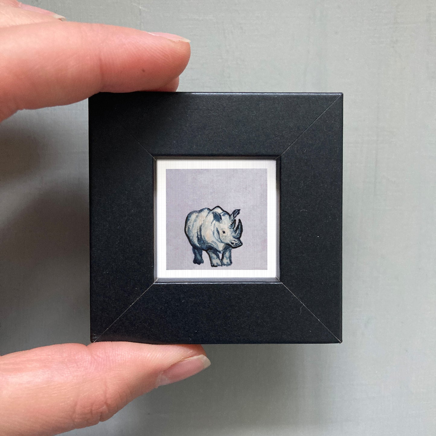A person's hand is holding a small black square frame containing a small illustration of a rhinoceros.
