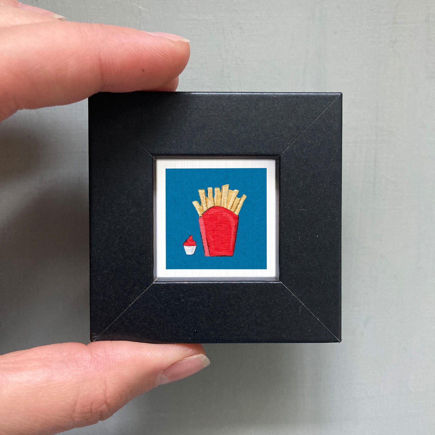 A hand is holding a small black frame containing a framed image of a red cup with french fries inside.