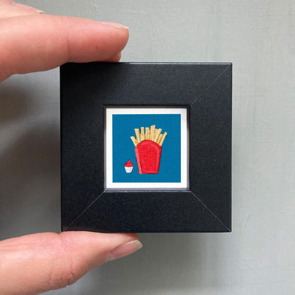 A hand is holding a small black frame containing a framed image of a red cup with french fries inside.