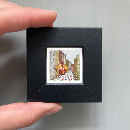 A person's hand is holding a small, square-shaped black frame containing a miniature painting of a city street scene.