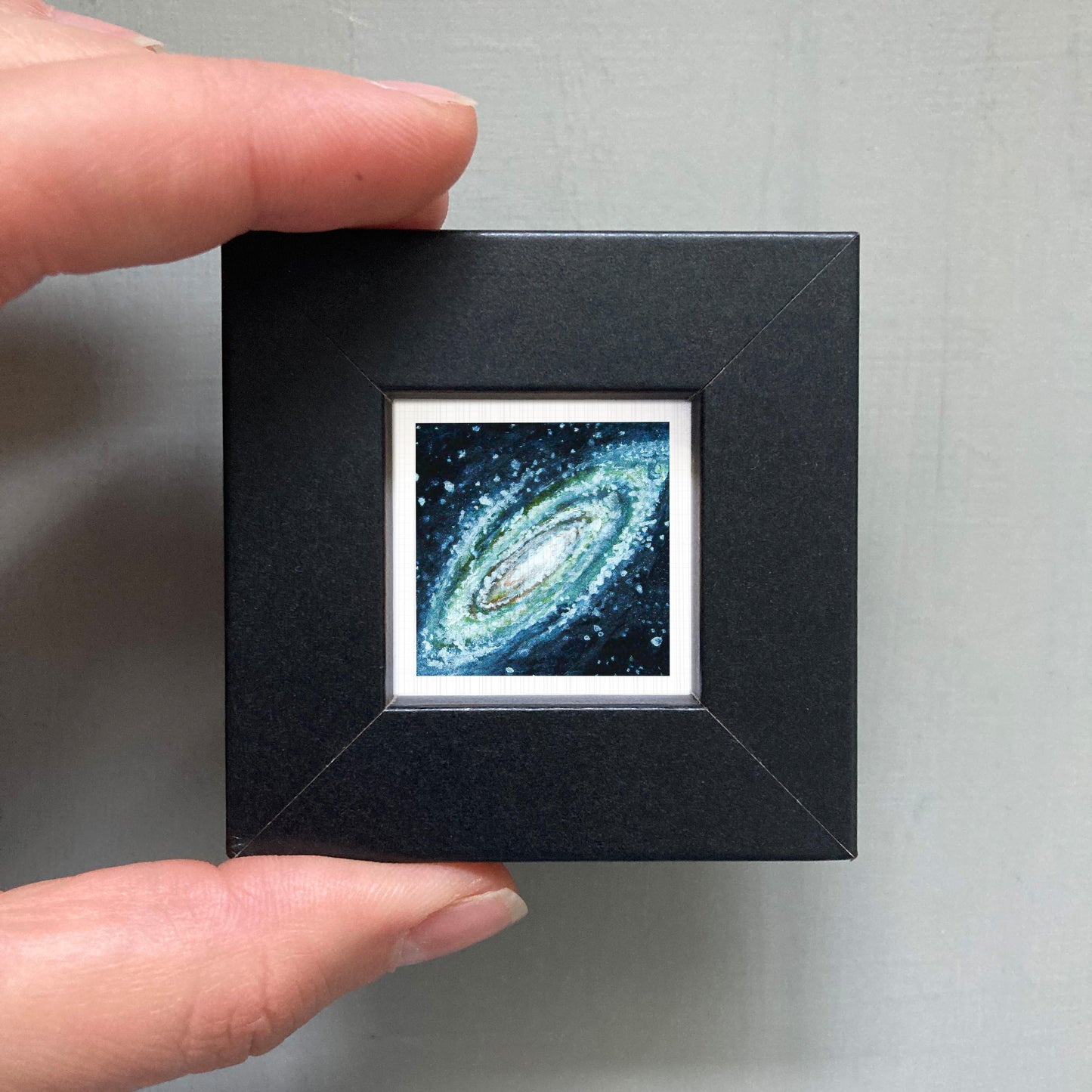 A person's hand is holding a small black frame containing a framed image of a spiral galaxy.