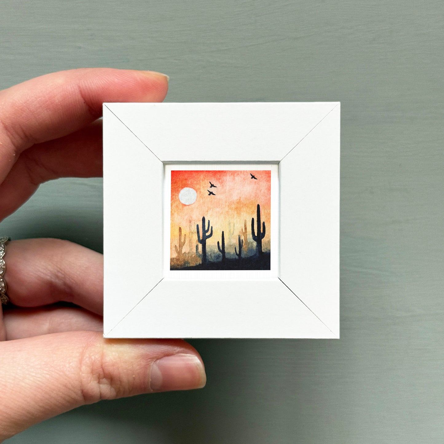 A hand is holding a small white square frame containing a painting of cacti against a sunset sky.