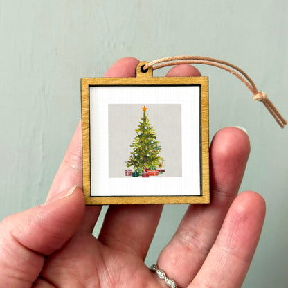 A hand holding a small wooden frame containing a picture of a Christmas tree.