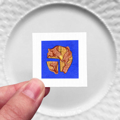A hand is holding a small square with a colorful illustration of a piece of food, such as a slice of pizza or a piece of cake, against a white background.