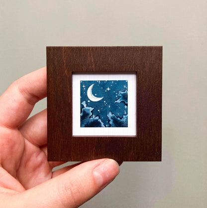 A hand is holding a small square picture frame containing a night sky scene with a crescent moon and stars.