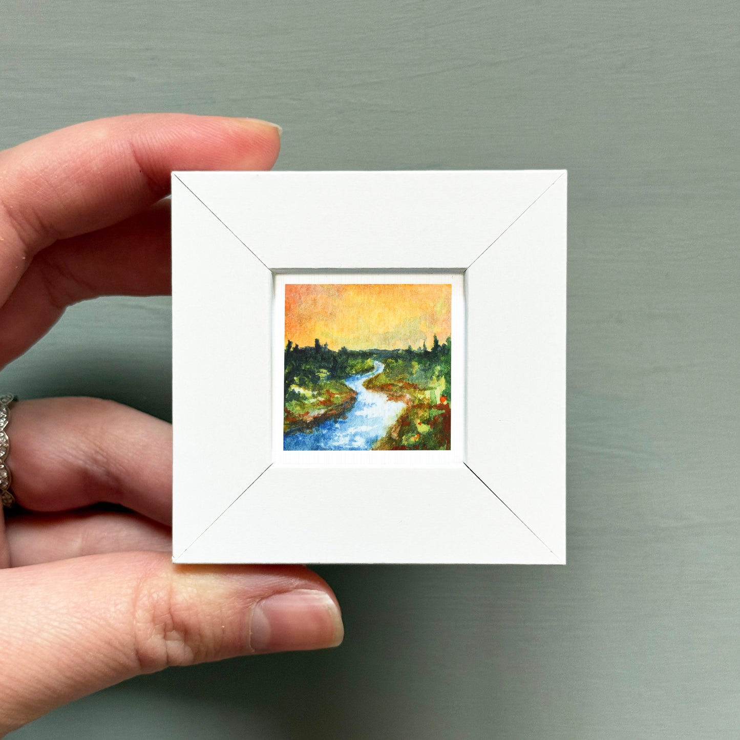 A hand is holding a small white square frame containing a painting of a river and trees.