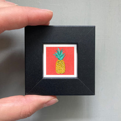 A hand is holding a small black frame containing a vibrant red square with a pineapple illustration on it.