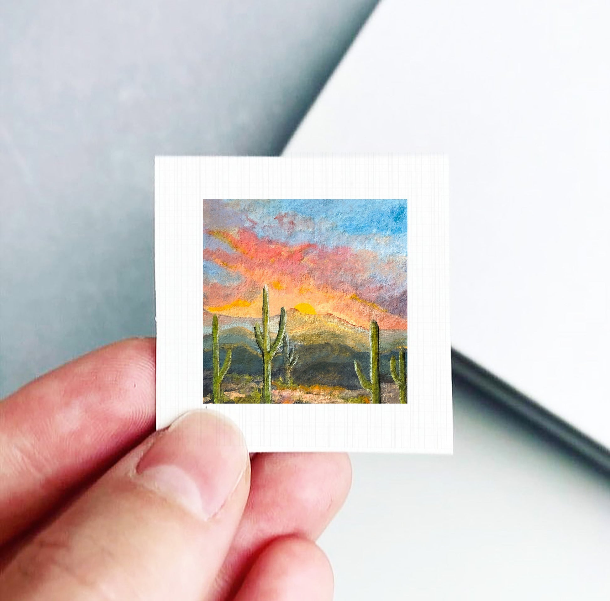 A hand is holding a small square picture of a desert landscape with cacti and a colorful sunset sky.