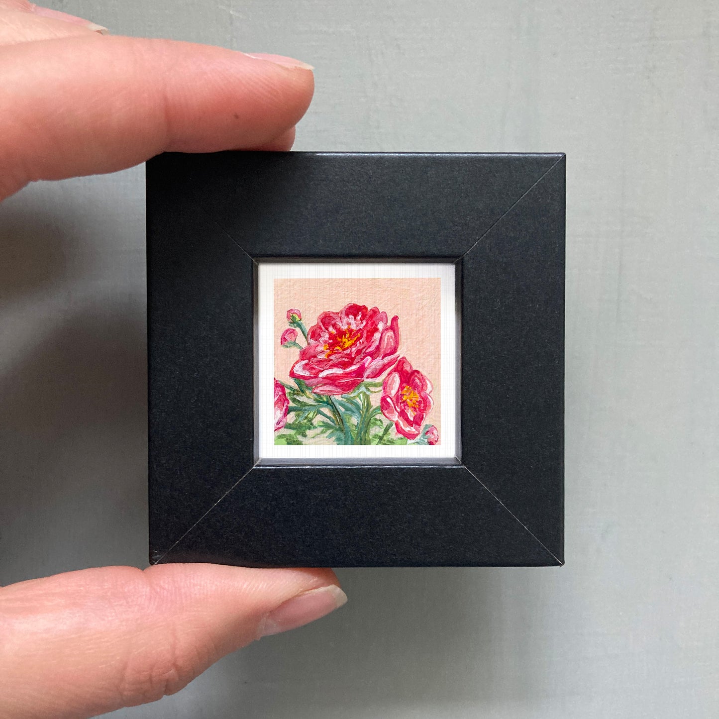 A person's hand holding a small black frame containing a framed painting of two pink flowers.