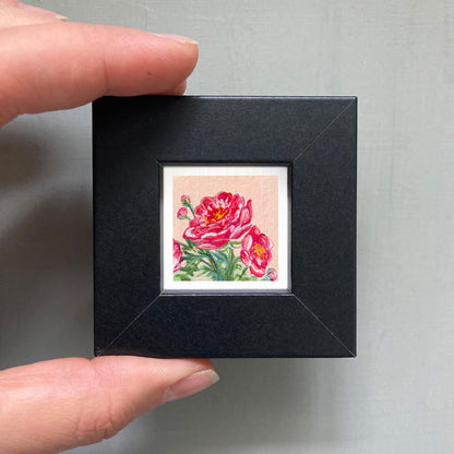 A person's hand holding a small black frame containing a framed painting of two pink flowers.