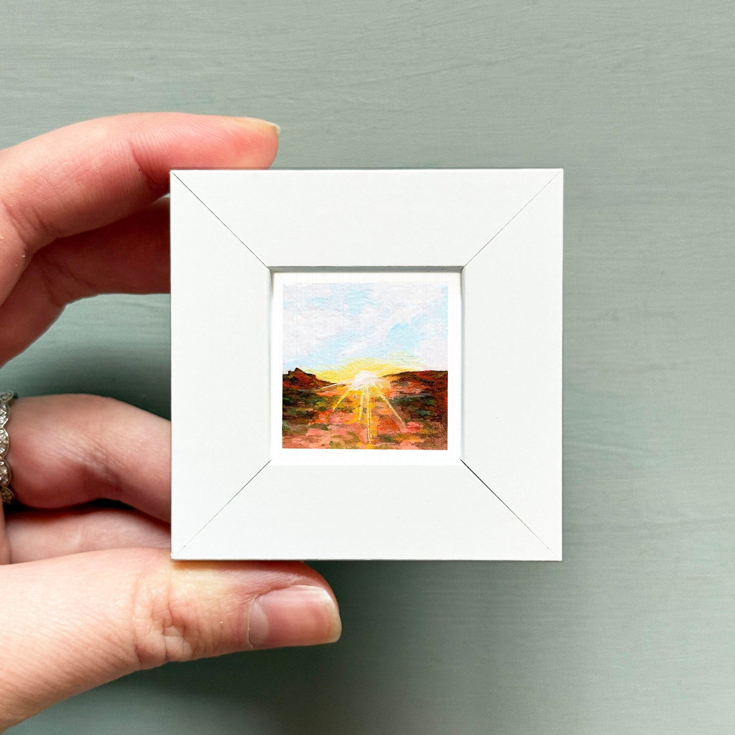 A hand is holding a small white square frame containing a painting of a landscape with a sunset.