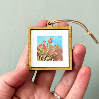 A hand holding a small wooden frame containing a framed painting of red flowers against a blue sky.