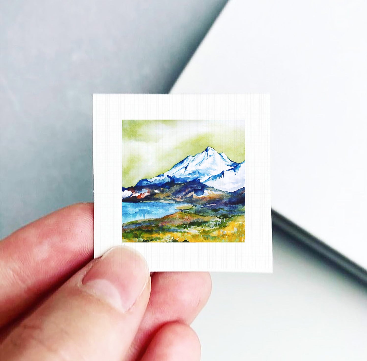 A hand is holding a small square artwork depicting a mountain landscape with a lake and green grass.