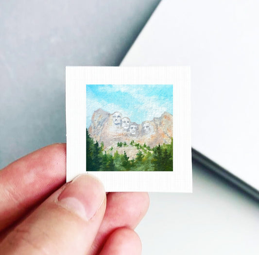 A person's hand is holding a small square picture depicting the iconic Mount Rushmore against a backdrop of a blue sky and green trees.