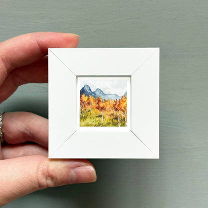 A hand is holding a small white frame containing a watercolor painting of a mountain landscape with autumn trees.