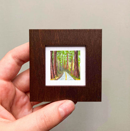 A hand is holding a small wooden frame containing a painting of a forest path with tall trees.