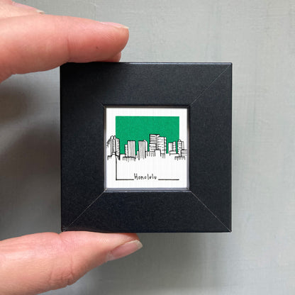 A hand is holding a small black square frame containing a drawing of a city skyline with green buildings.