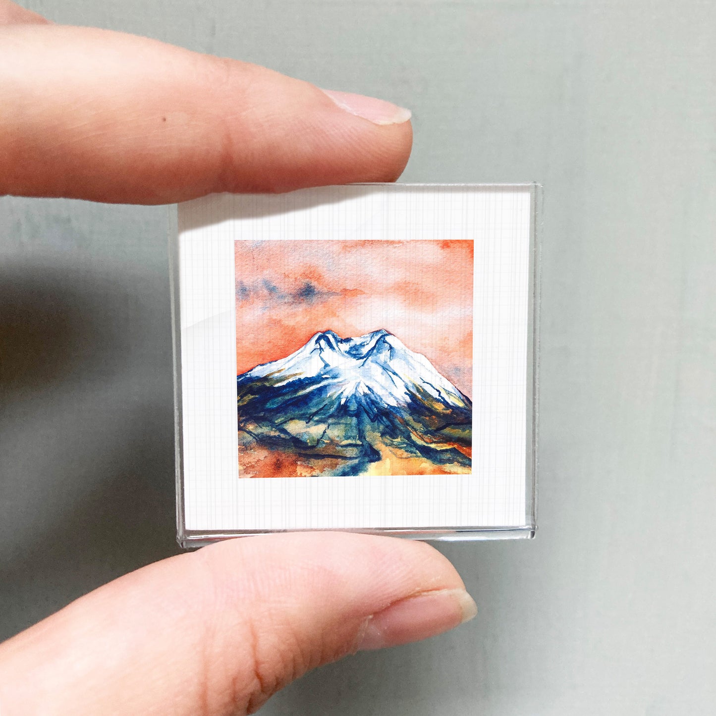 A person's hand holding a small square picture of a mountain landscape with a watercolor painting of the mountain.