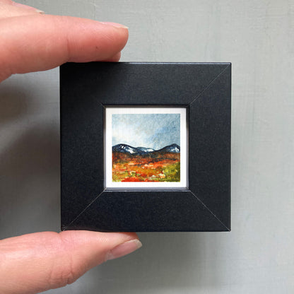 A person's hand holding a small, square-shaped black frame containing a watercolor painting of a mountain landscape.