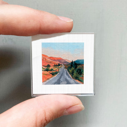 A person's hand holding a small square picture of a scenic landscape with a winding road and mountains in the background.