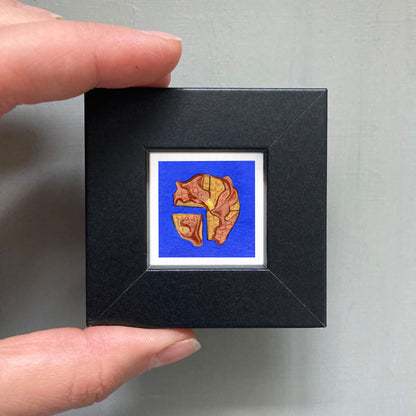 A person's hand is holding a small square-shaped black frame containing a vibrant, colorful illustration of a heart shape.